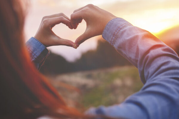 Two friends forming a heart shape with their hands against a golden sunset sky